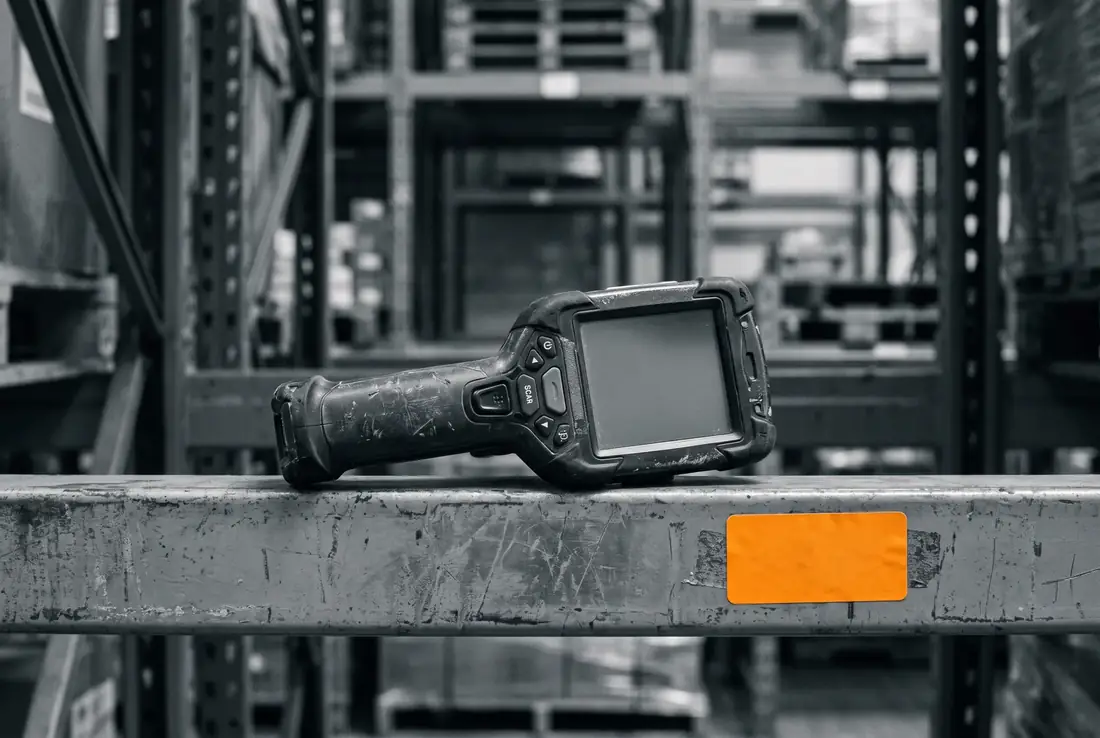 A worn, scuffed handheld rugged barcode scanner resting on a warehouse racking beam, next to a blank orange shipping label.